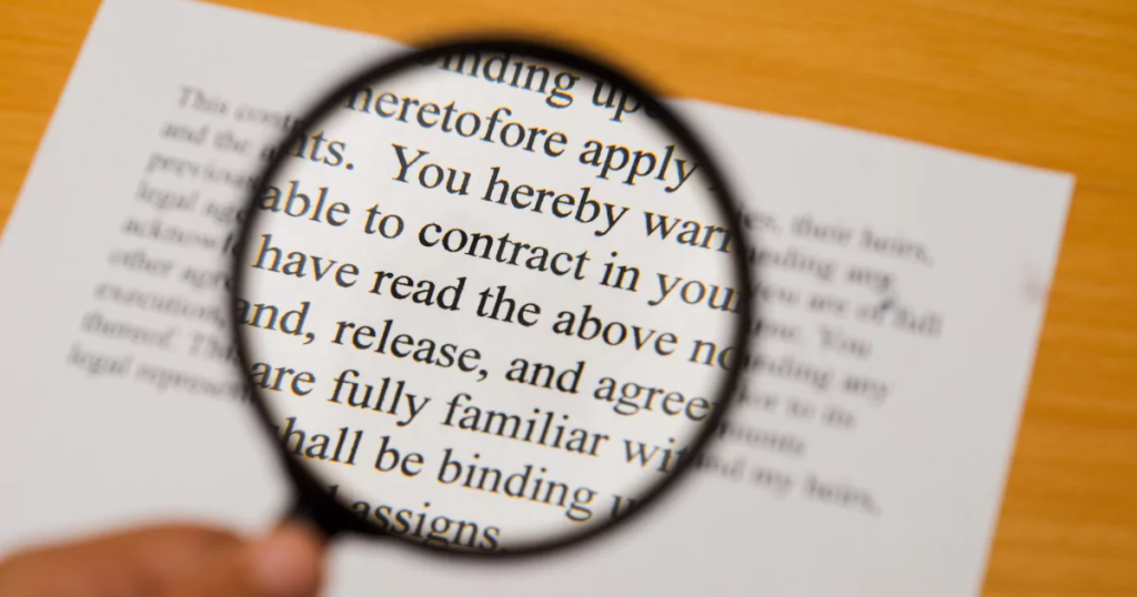 Primer plano de una mano sosteniendo una lupa negra sobre un contrato legal impreso, ampliando el texto para leer detenidamente las cláusulas y condiciones antes de aceptarlas.