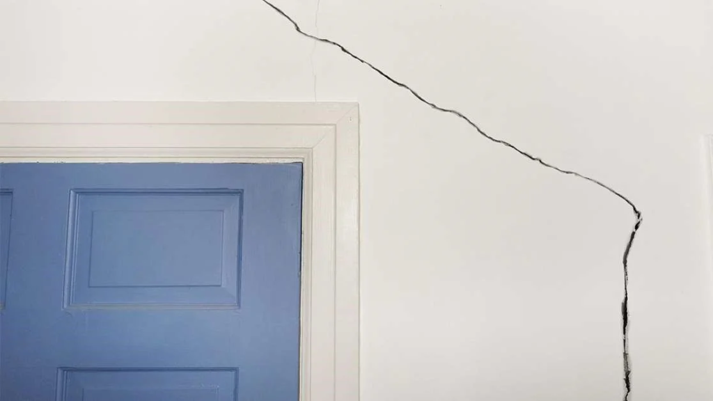 Grieta diagonal pronunciada en pared blanca junto a marco de puerta azul