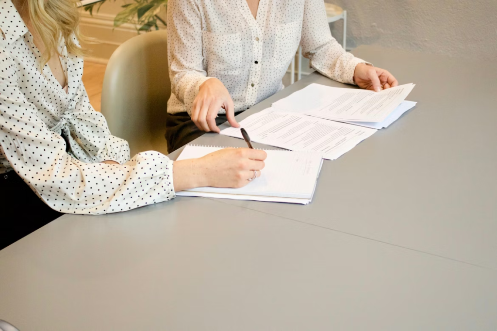 Dos mujeres sentadas frente a una mesa gris y lisa revisando detenidamente unos contratos; una de ellas sostiene un bolígrafo y está a punto de firmar o tomar notas en una libreta.