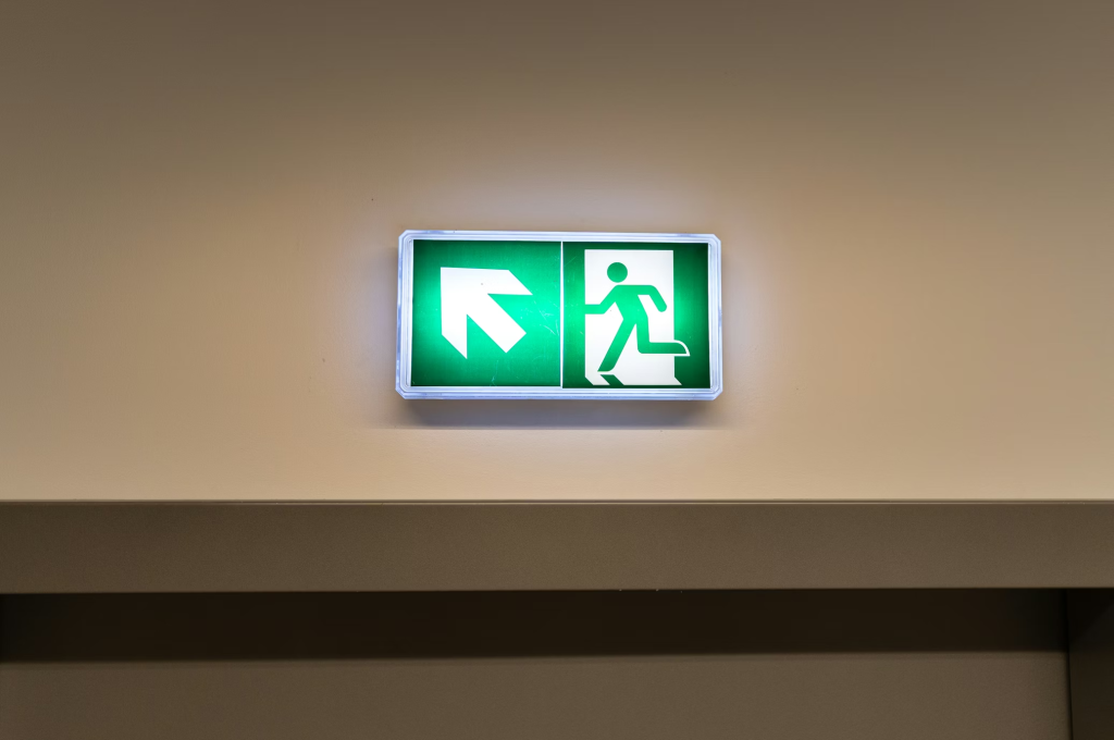 Letrero luminoso verde de salida de emergencia montado en una pared, que muestra la silueta blanca de una persona corriendo hacia la salida junto a una flecha.