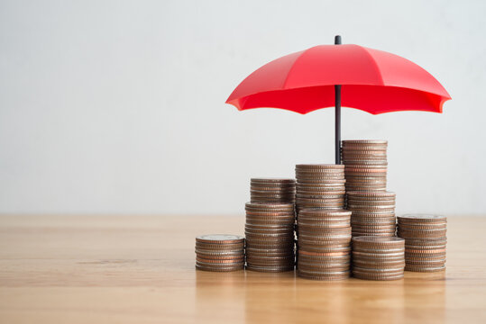 Varias columnas de monedas apiladas sobre una mesa de madera, cobijadas y protegidas bajo un pequeño paraguas rojo abierto sobre un fondo gris claro.