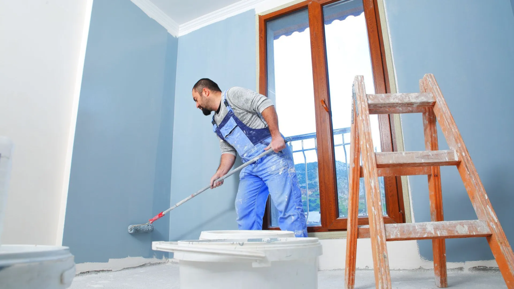 Operario con mono azul aplicando pintura en el suelo de una habitación durante reforma de vivienda