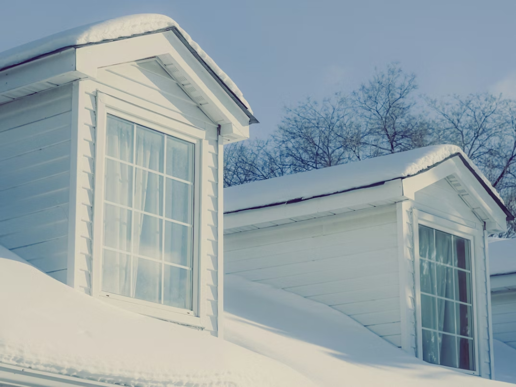 Tejado de una casa de madera blanca cubierto por una gruesa capa de nieve con dos ventanas tipo buhardilla, ilustrando la exposición al clima invernal.