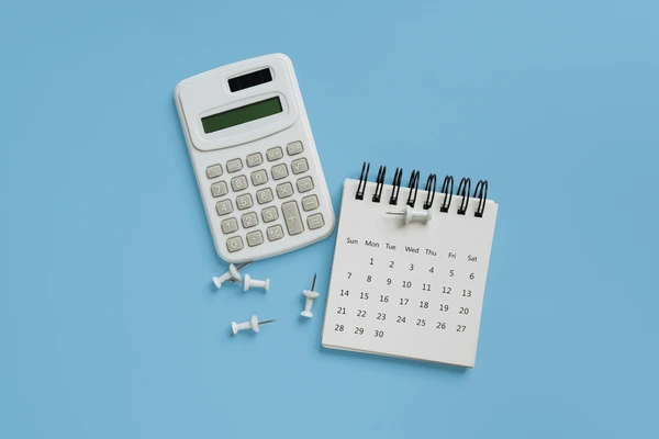 Calculadora blanca junto a un pequeño calendario de anillas y unas chinchetas sobre un fondo azul claro, representando el cálculo de fechas y números en las finanzas personales.