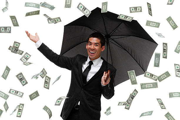 Hombre de negocios sonriente protegiéndose con un paraguas negro abierto bajo una lluvia de billetes, simbolizando la liquidez financiera y la protección ante imprevistos.