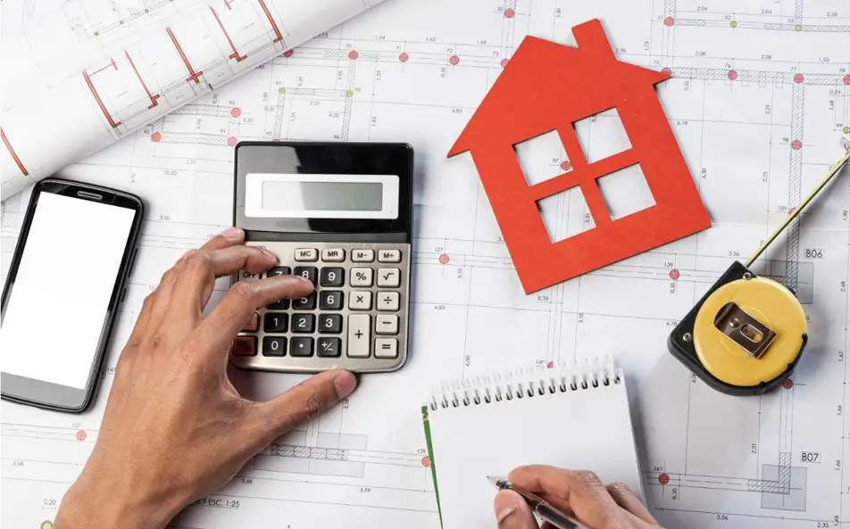Manos usando calculadora sobre planos de obra con figura de casa roja y cinta métrica