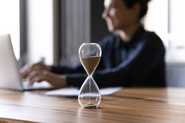 Reloj de arena en primer plano sobre mesa con persona trabajando al ordenador al fondo