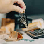 Mano sosteniendo llave de casa con llavero en forma de vivienda junto a billetes y calculadora