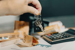 Mano sosteniendo llave de casa con llavero en forma de vivienda junto a billetes y calculadora