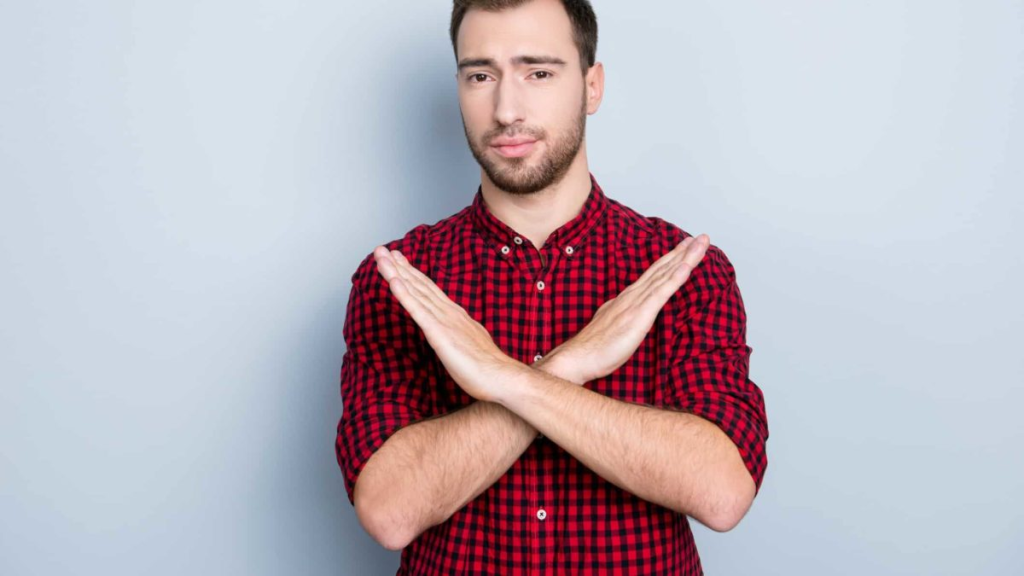 Hombre joven con camisa roja haciendo gesto de negación con los brazos cruzados sobre fondo gris