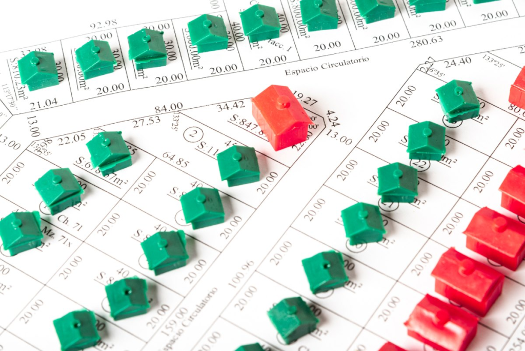 Pequeñas casas de plástico de colores verde y rojo esparcidas sobre un plano catastral o mapa urbanístico impreso en papel con divisiones y medidas topográficas.