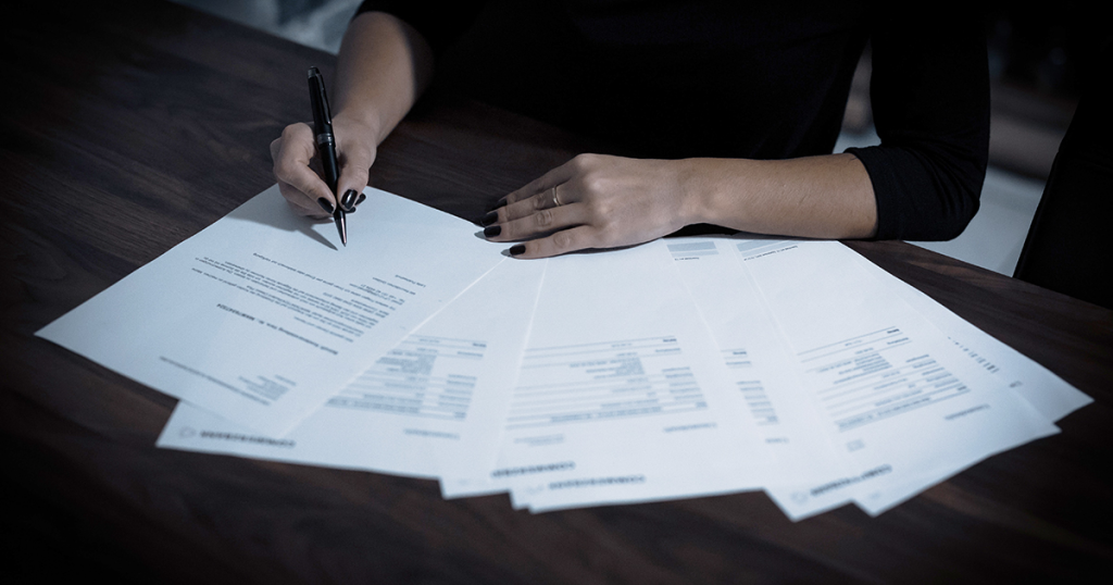 Mujer firmando varios documentos extendidos sobre mesa oscura en proceso de compraventa inmobiliaria