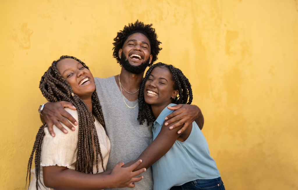 Tres personas jóvenes abrazadas y sonriendo frente a pared amarilla en actitud celebratoria