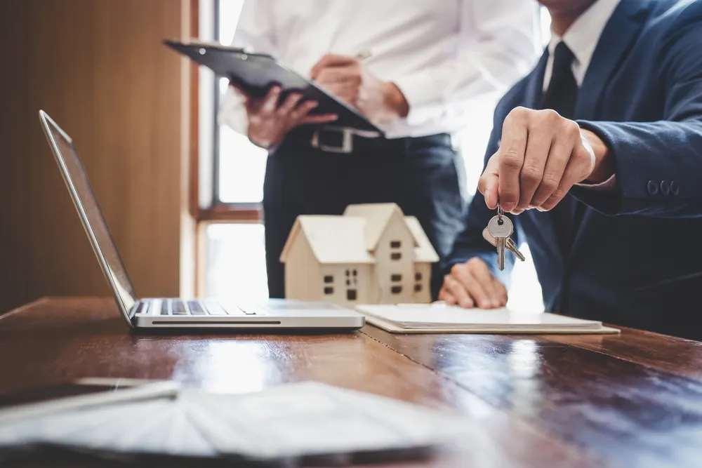 Agente inmobiliario vestido con traje entregando unas llaves hacia la cámara sobre un escritorio de madera, donde hay un modelo de casa, un ordenador portátil y documentos.