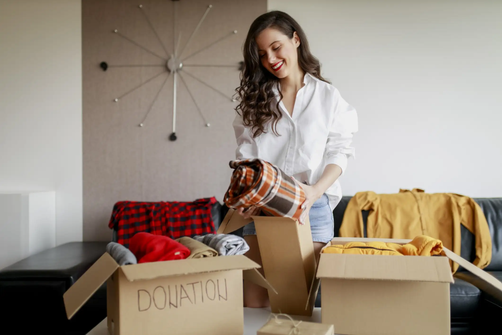 Mujer joven sonriente empaquetando ropa en caja de donación en salón antes de la mudanza