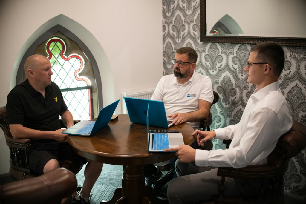 Tres hombres reunidos alrededor de mesa redonda con portátiles en sala con ventanales de estilo gótico