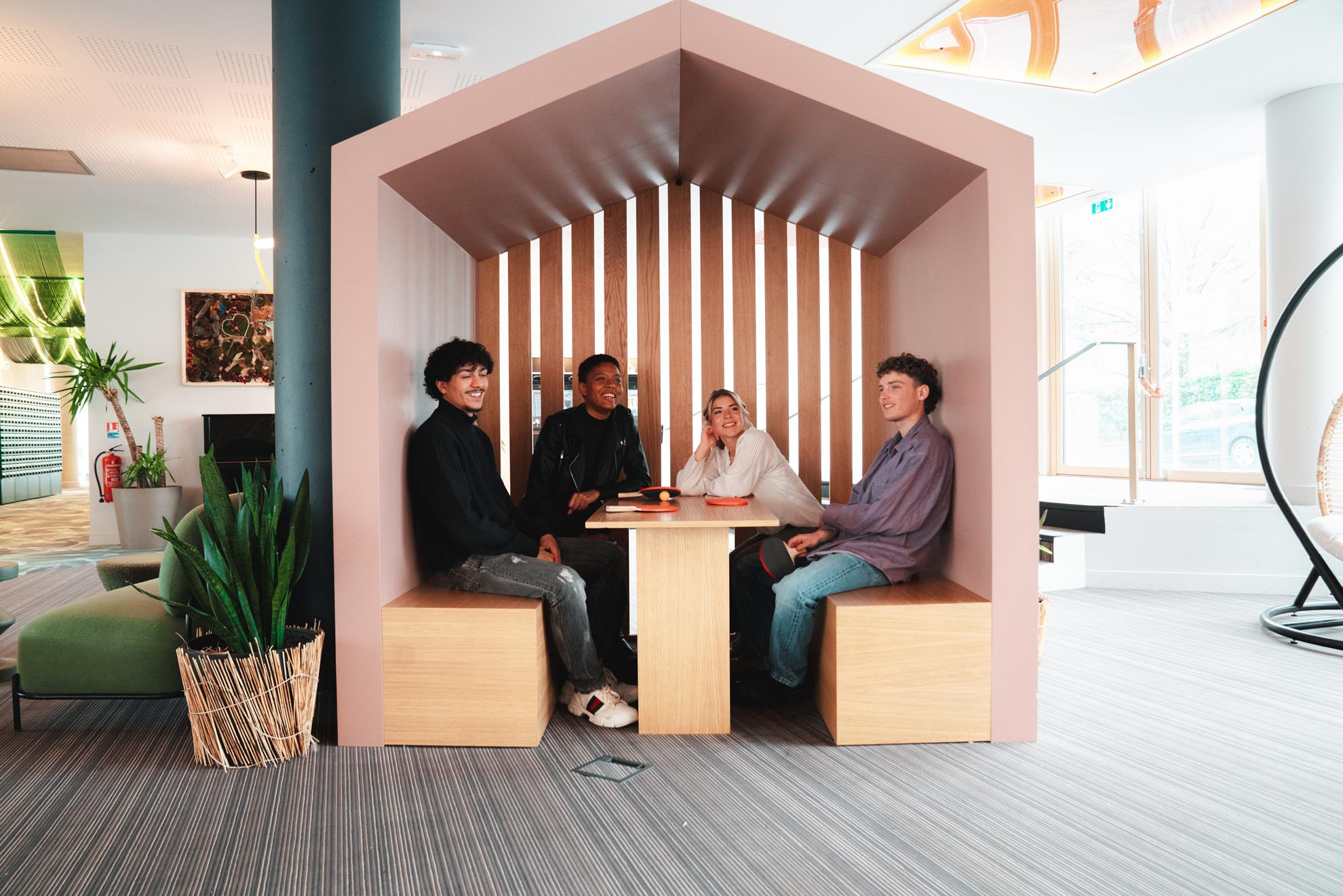 Grupo de cuatro jóvenes diversos sonriendo y charlando animadamente mientras están sentados en una moderna estructura de madera con forma de casa dentro de un espacio compartido.