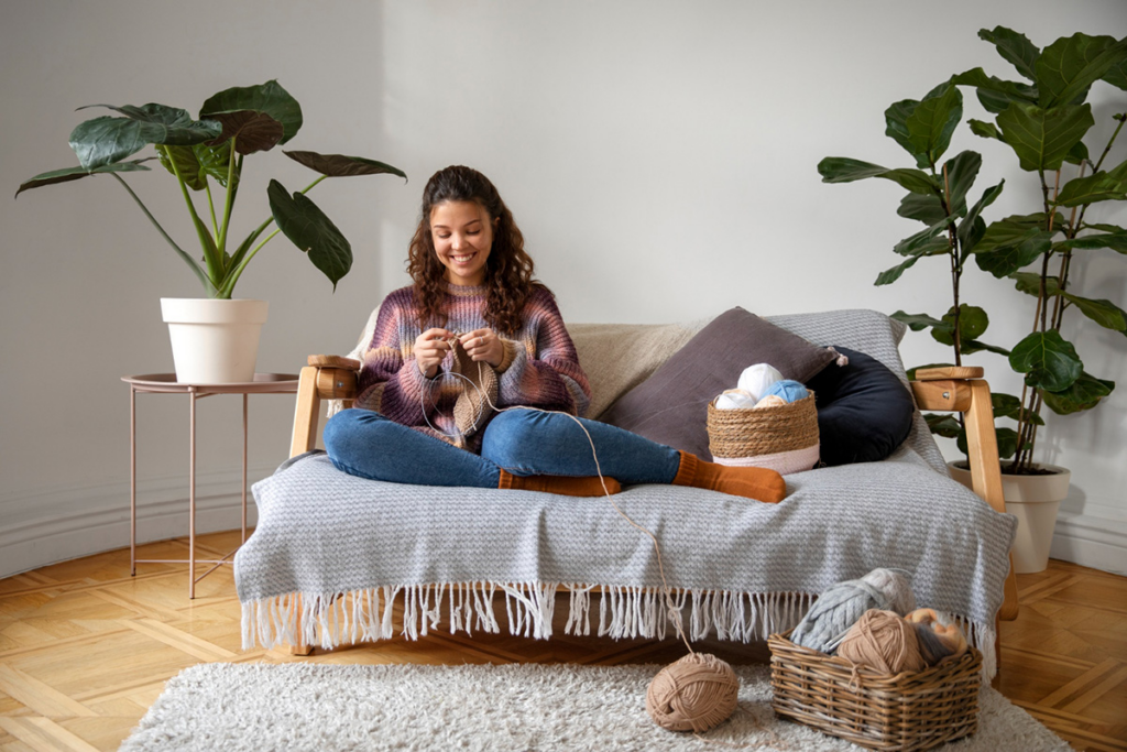 Mujer joven sonriendo y tejiendo relajadamente mientras está sentada con las piernas cruzadas en el sofá de su acogedor salón, decorado con plantas de interior y suelo de madera.