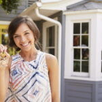 Mujer joven sonriendo frente a una casa, sosteniendo unas llaves en la mano, simbolizando la compra o adquisición de una vivienda.
